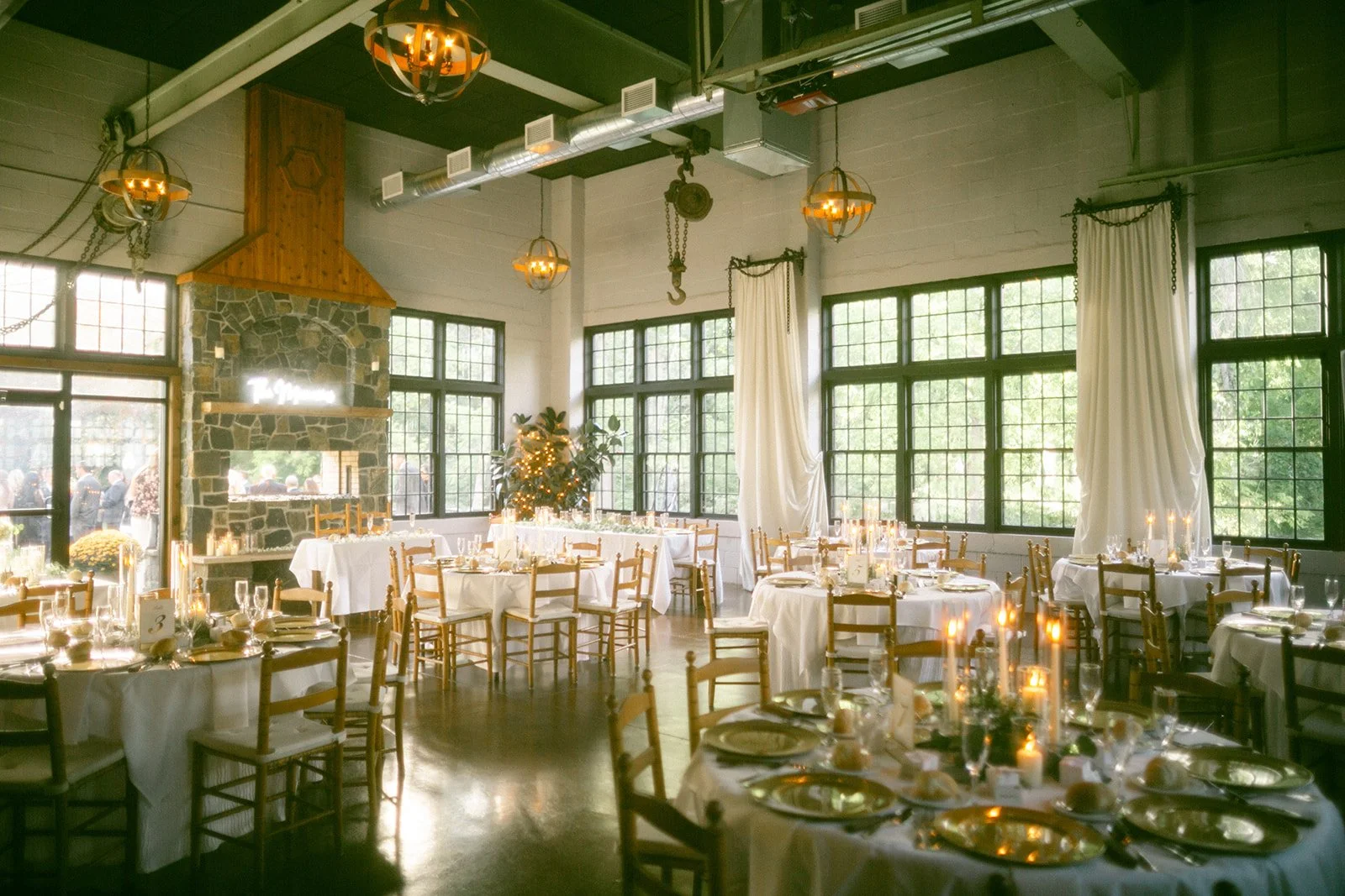 Sunlit dining room set for an event with round tables, wooden chairs, white tablecloths, candles, and large windows. Upstate New York Wedding Venue