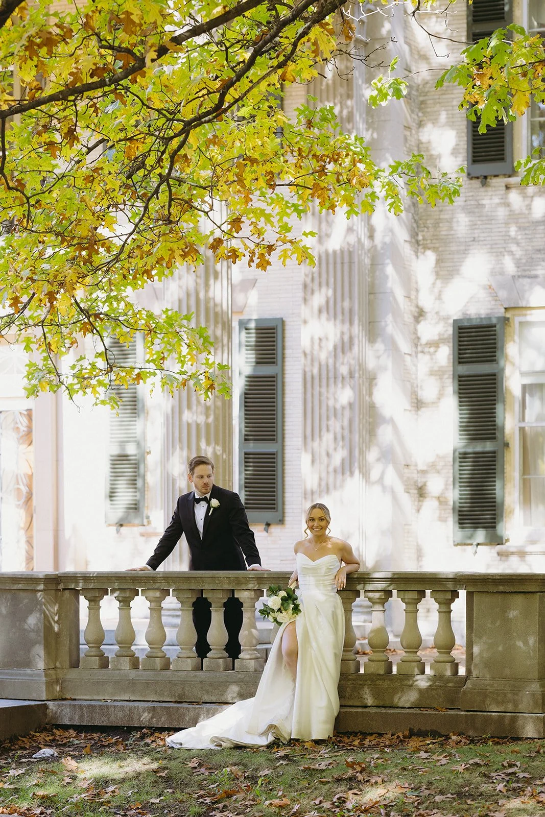 A bride and groom in formal wedding attire walk down stone steps outside a large historic building with columns at George Eastman Museum an upstate new york wedding venue 
