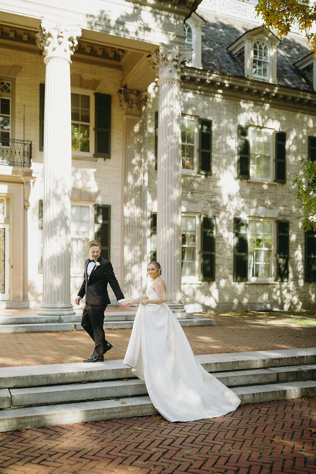 A bride and groom in formal wedding attire walk down stone steps outside a large historic building with columns at George Eastman Museum an upstate new york wedding venue 