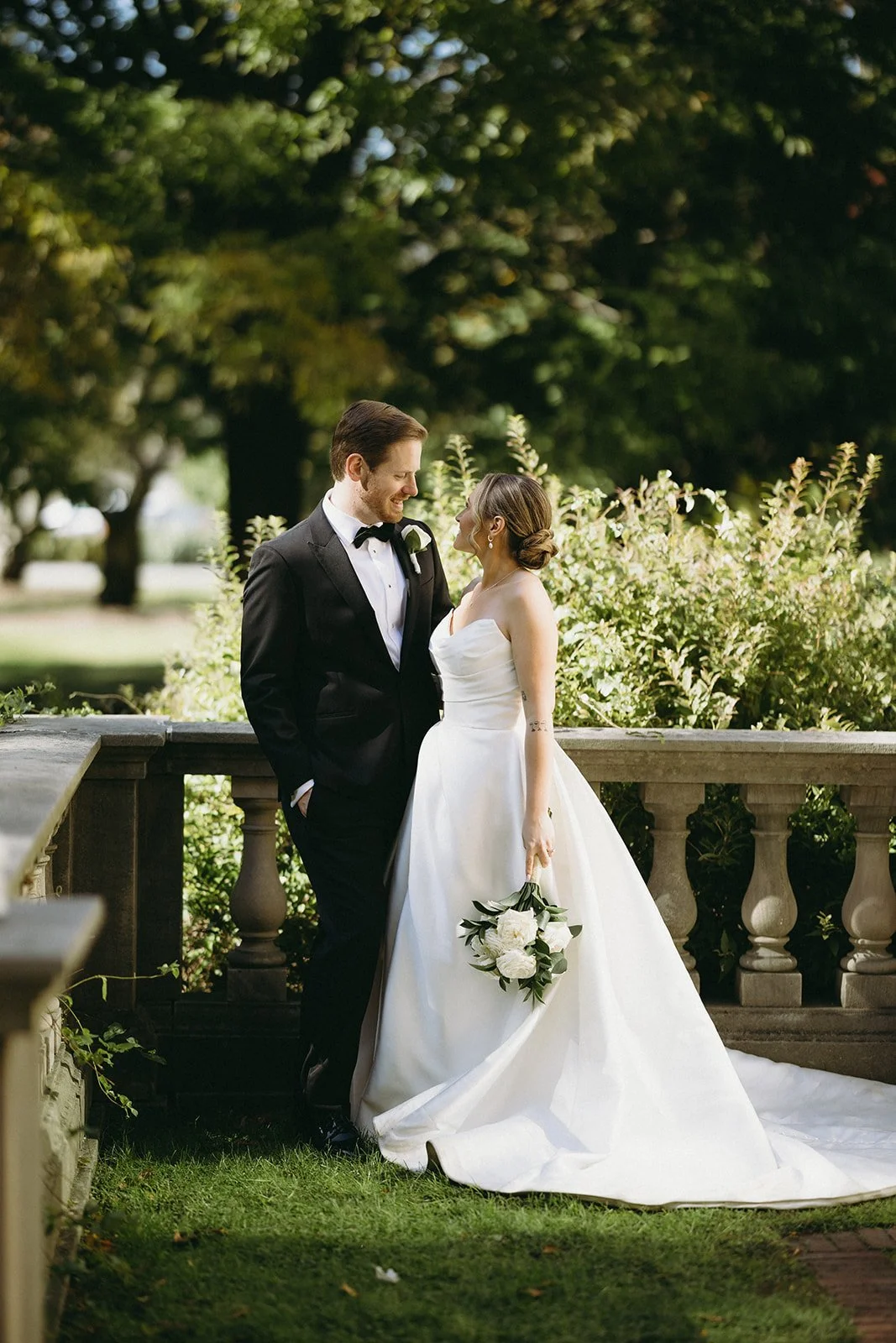A bride and groom stand together outside a large building with tall columns, surrounded by trees and sunlight.