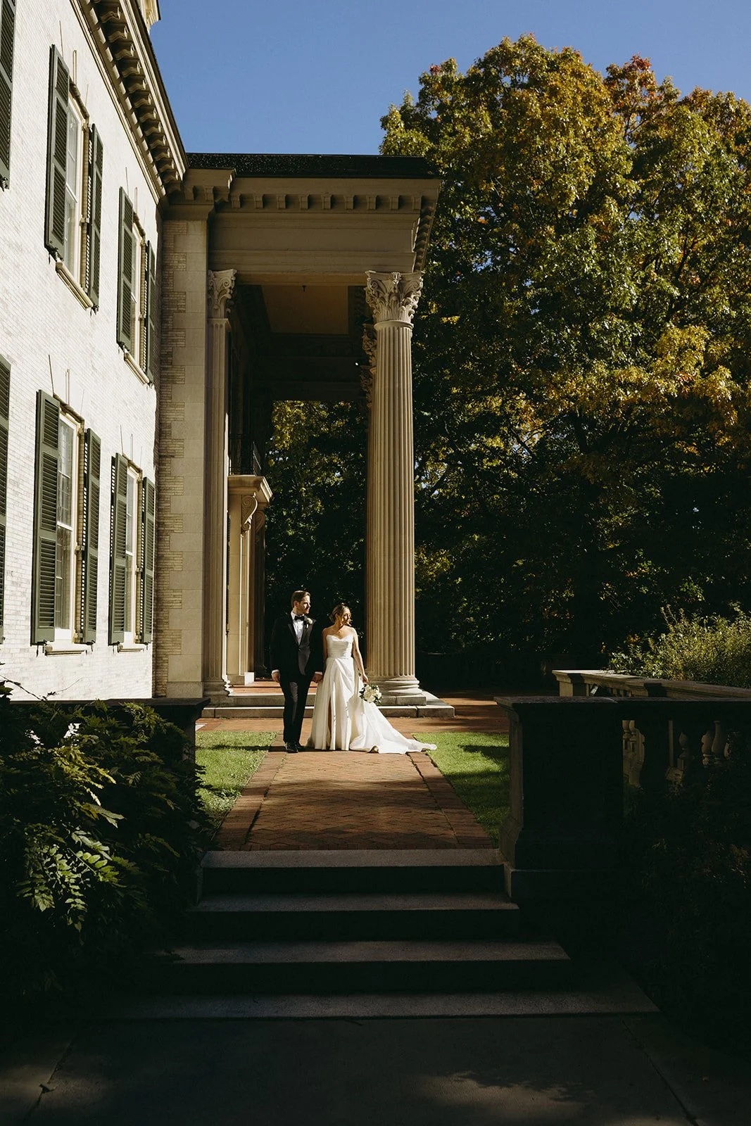 A bride and groom stand together outside a large building with tall columns, surrounded by trees and sunlight.