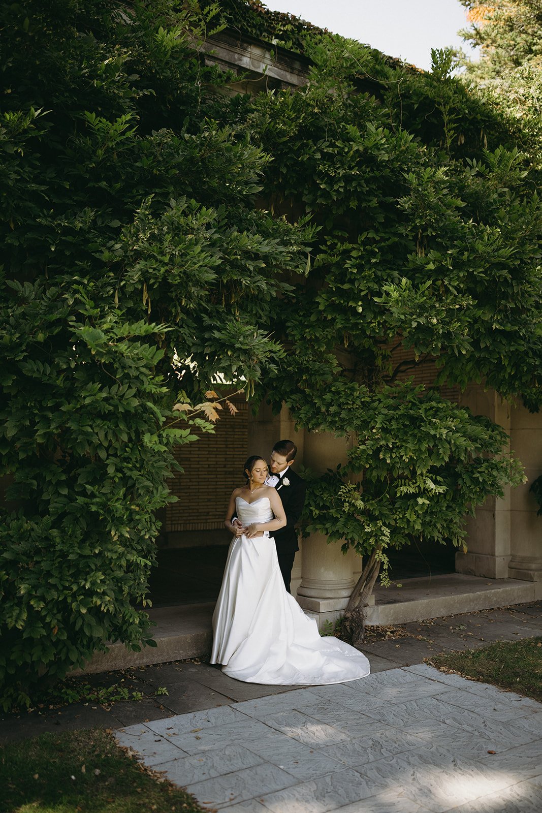 A bride in a white gown and a groom in a black suit stand together outside, surrounded by lush green foliage and stone architecture.
