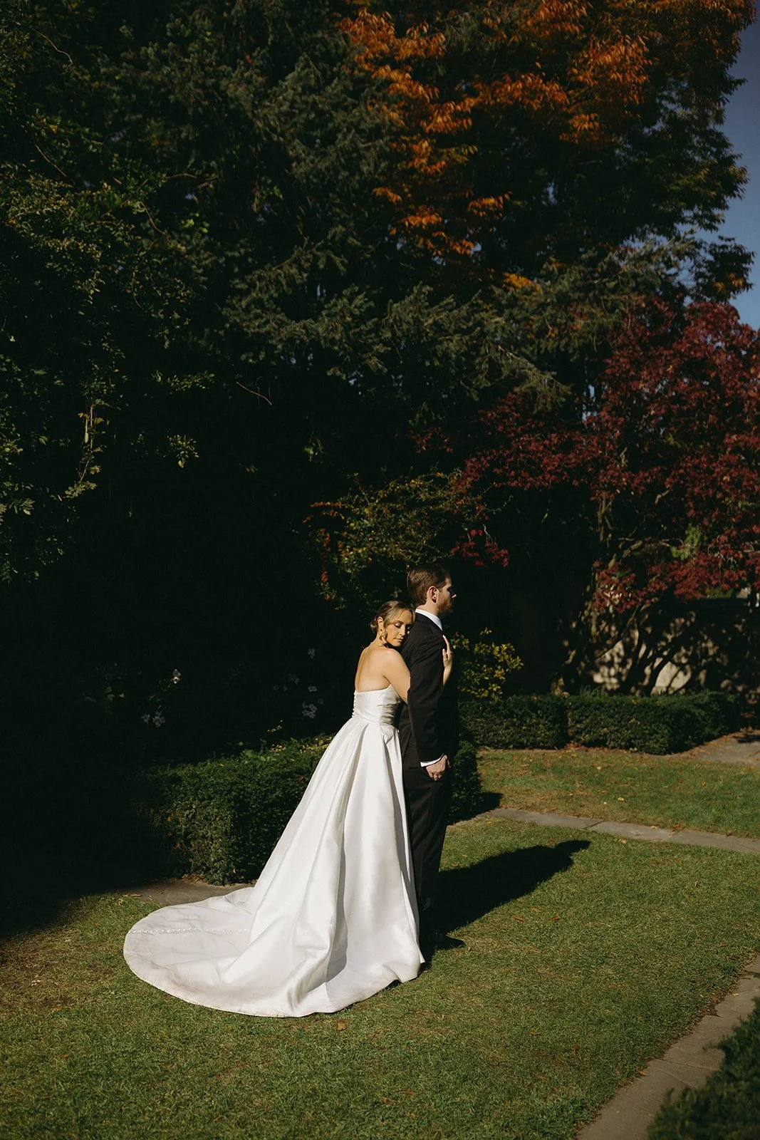 A bride in a white gown and a groom in a black suit stand together outside, surrounded by lush green foliage and stone architecture.