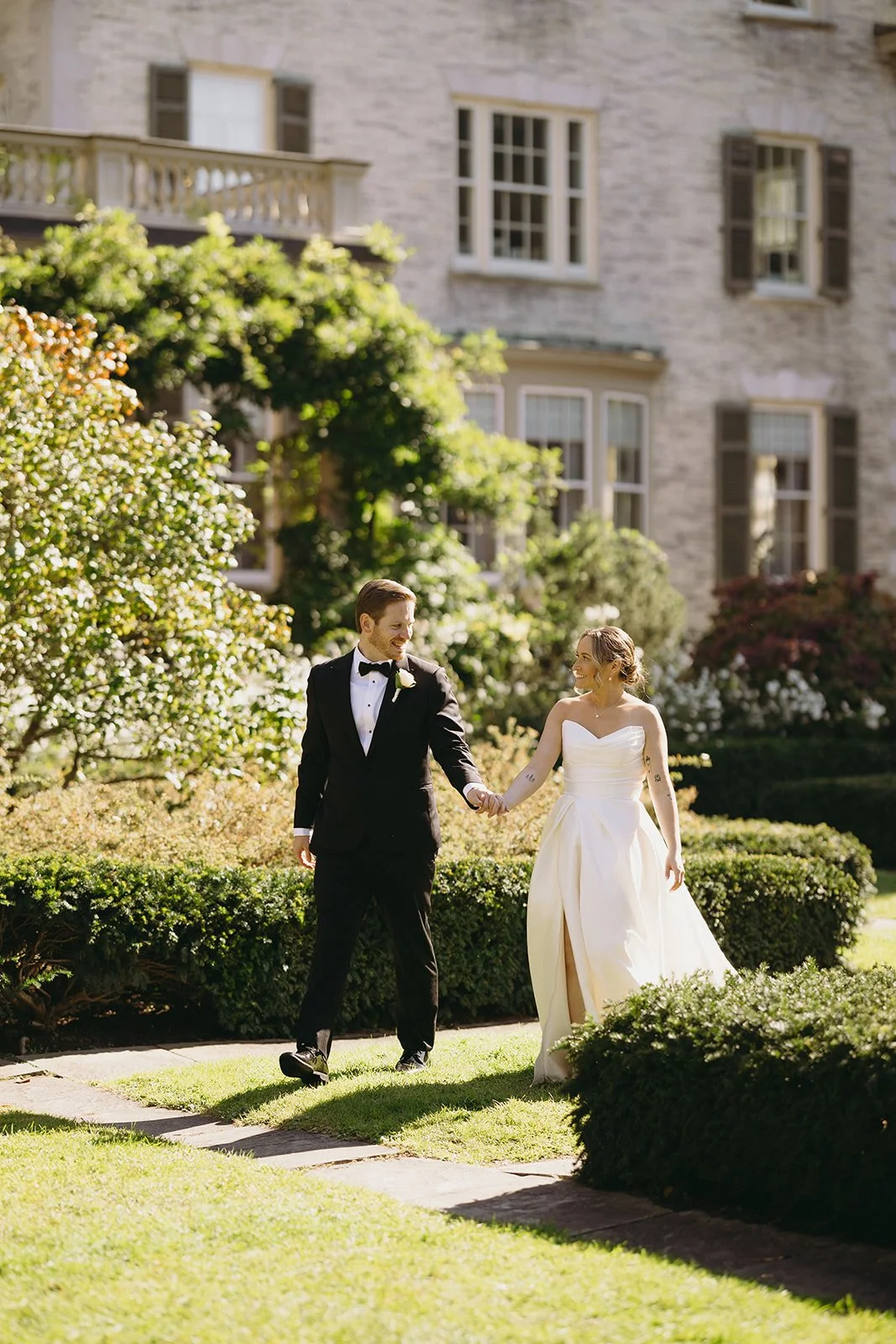 A bride in a white gown and a groom in a black suit stand together outside, surrounded by lush green foliage and stone architecture.