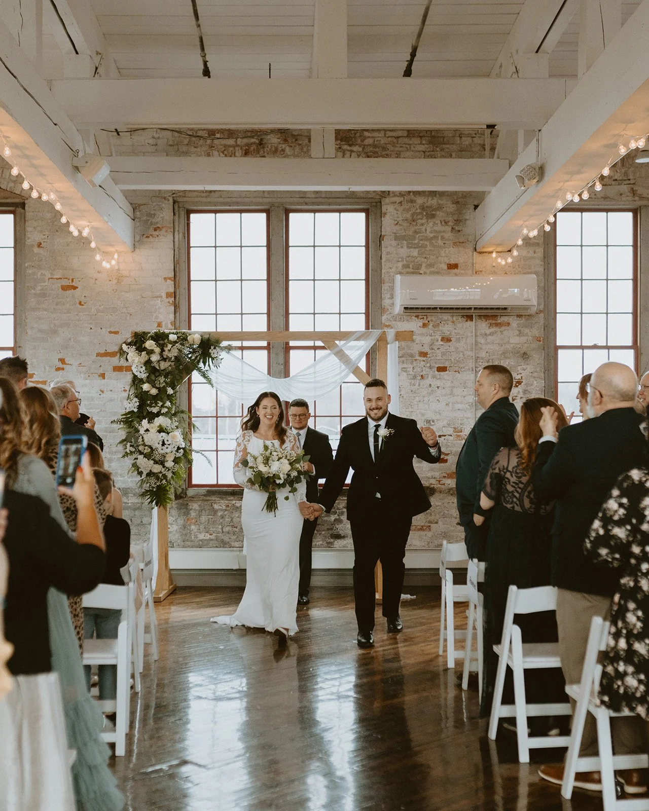 A bride and groom stand facing each other, holding hands during a wedding ceremony indoors with a floral arch and two officiants present.