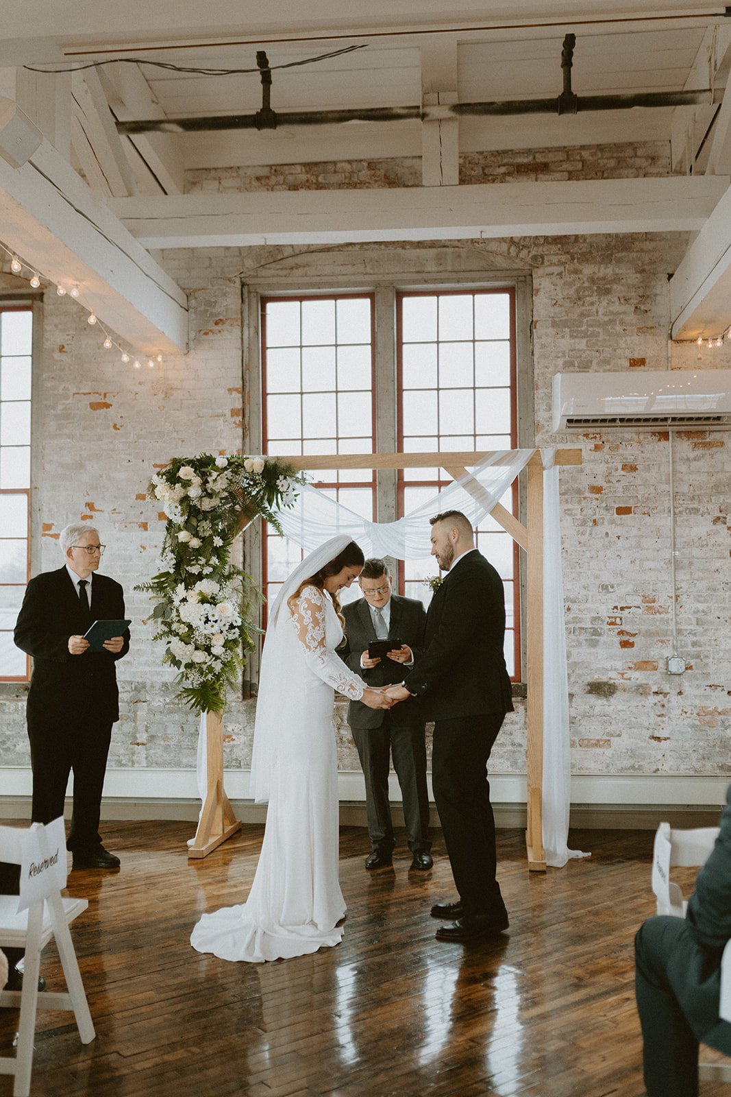 A bride and groom stand facing each other, holding hands during a wedding ceremony indoors with a floral arch and two officiants present.