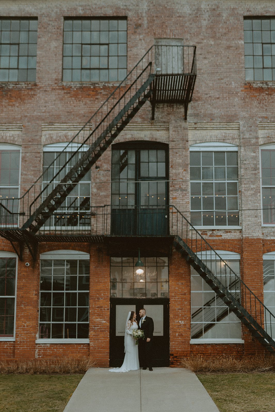 A bride and groom stand together in wedding attire, smiling in front of a brick building with glass doors and large windows.