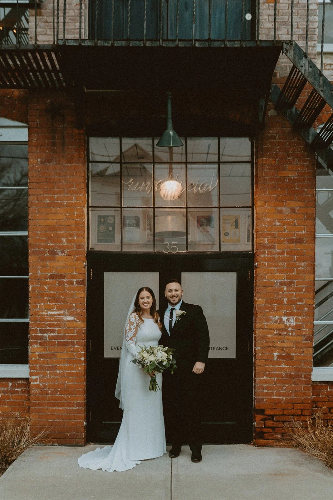 A bride and groom stand together in wedding attire, smiling in front of a brick building with glass doors and large windows.