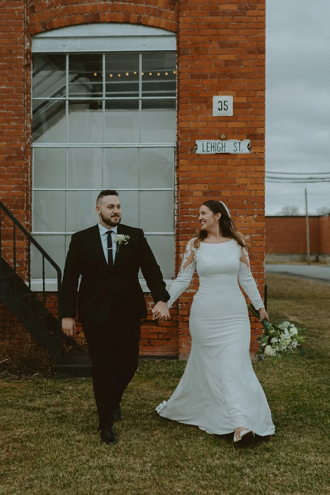 A bride and groom stand together in wedding attire, smiling in front of a brick building with glass doors and large windows.