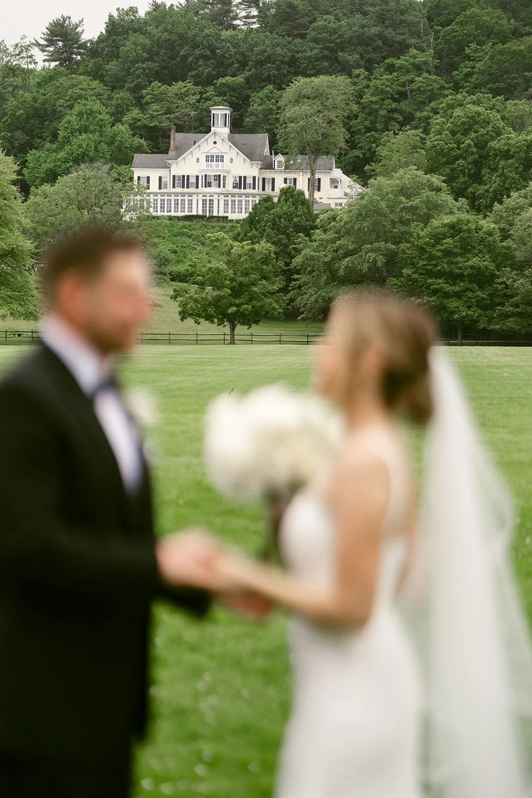 A bride and groom stand together on a grassy field, with the groom holding a bouquet and a large white house and trees visible in the background.