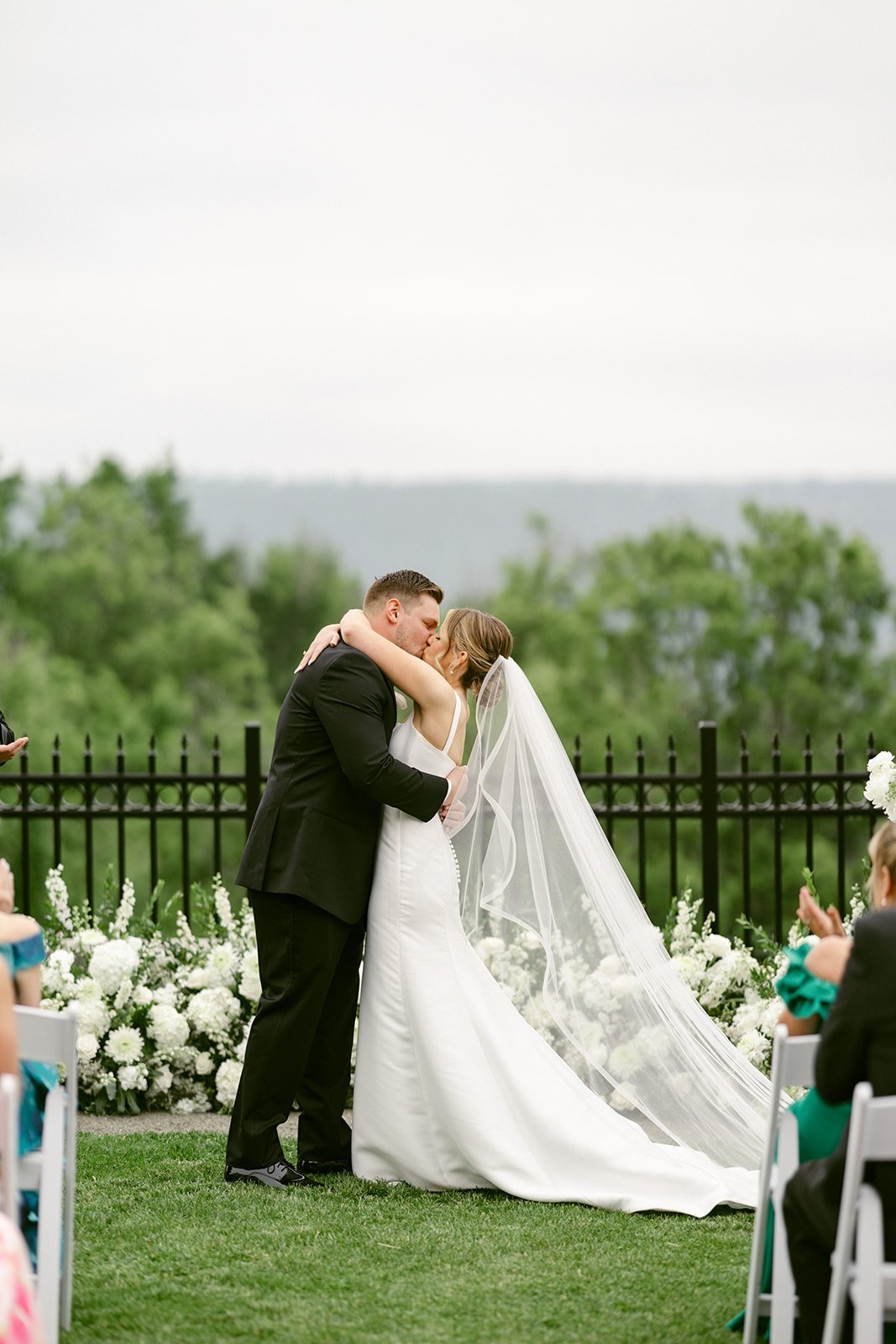 A bride and groom share a kiss at an outdoor wedding ceremony, standing in front of white flowers and a black fence, surrounded by guests.