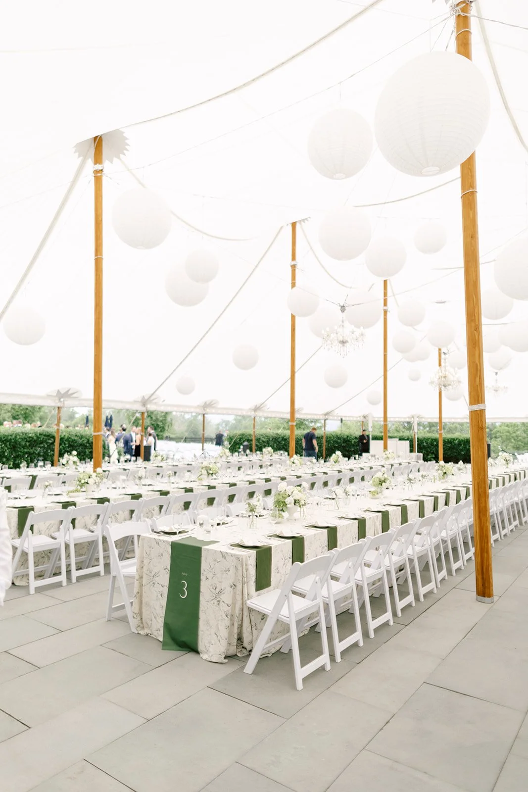 Rows of white tables with green runners and white chairs are arranged under a canopy with hanging white paper lanterns at an outdoor event.
