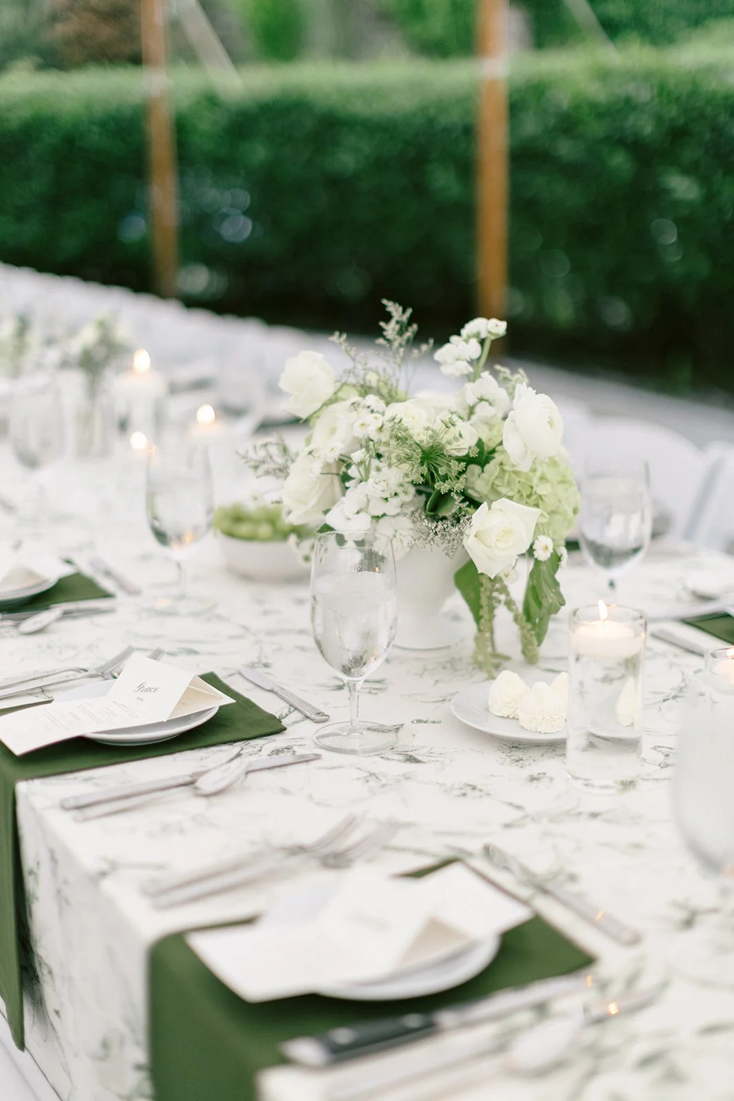 Rows of white tables with green runners and white chairs are arranged under a canopy with hanging white paper lanterns at an outdoor event.
