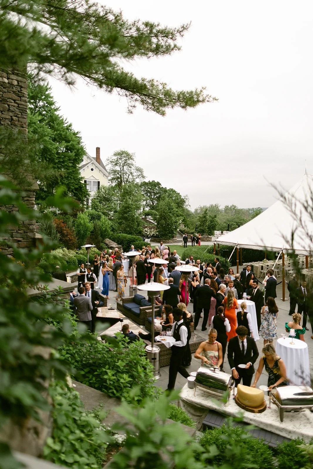 Outdoor formal event with guests in suits and dresses mingling around cocktail tables near a large tent, surrounded by greenery and stone structures.