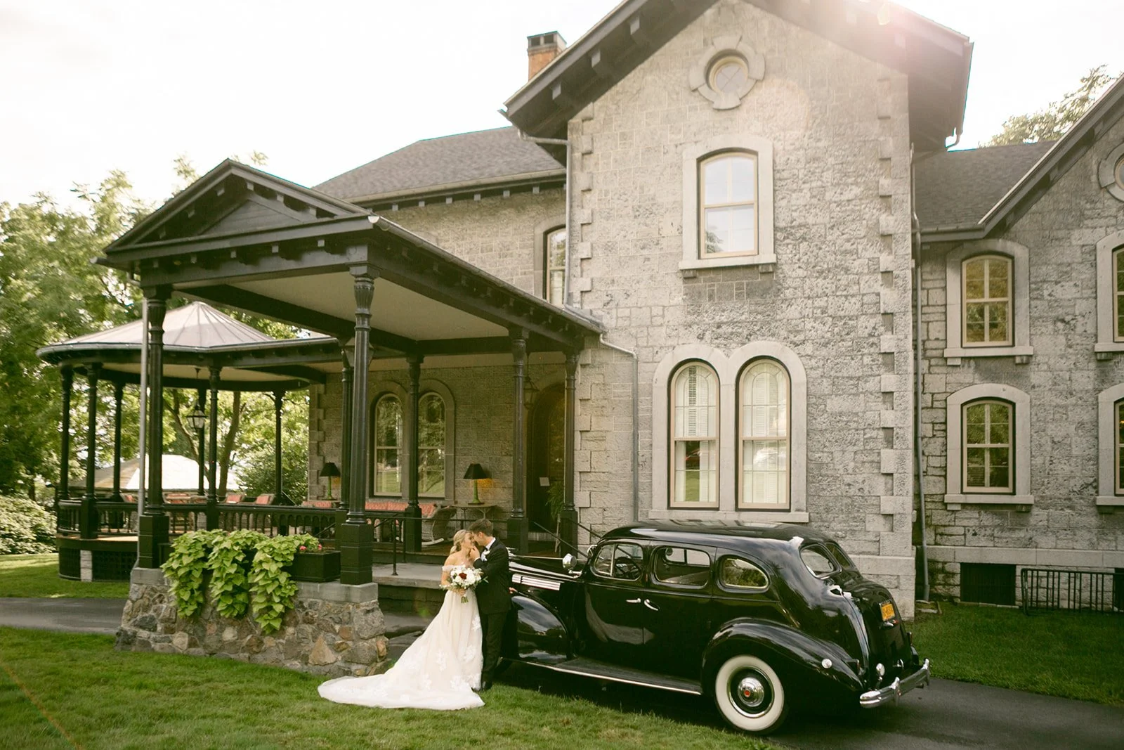 A bride and groom stand beside a vintage black car in front of a large stone house with arched windows and a covered porch at the inns of aurora an upstate ny wedding venue