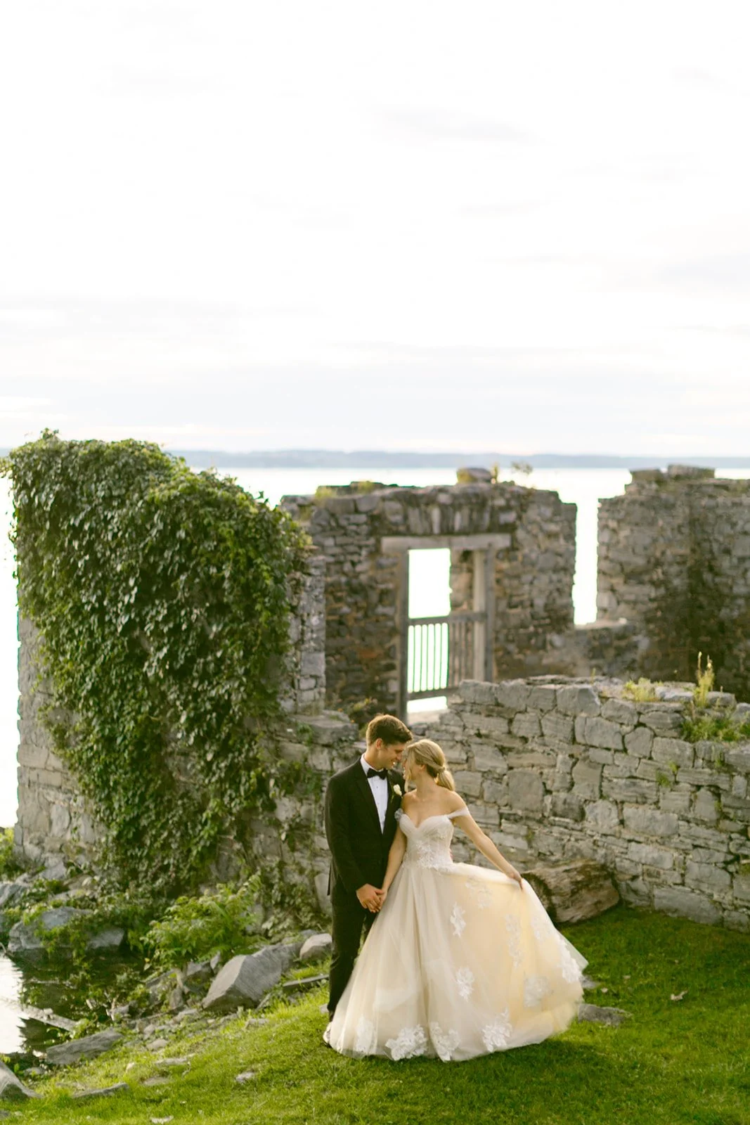 A bride and groom stand together in front of old stone ruins covered in ivy, with water visible in the background.
