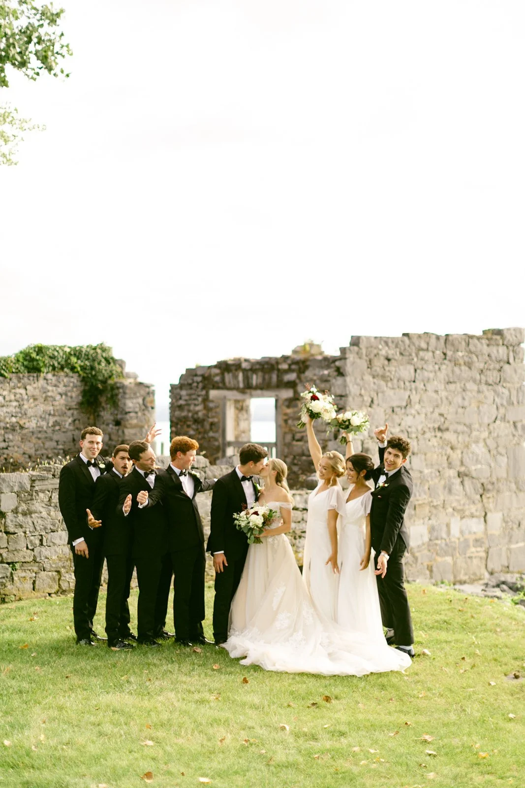 A wedding party of six people, dressed in formal attire, poses together on grass in front of old stone ruins.