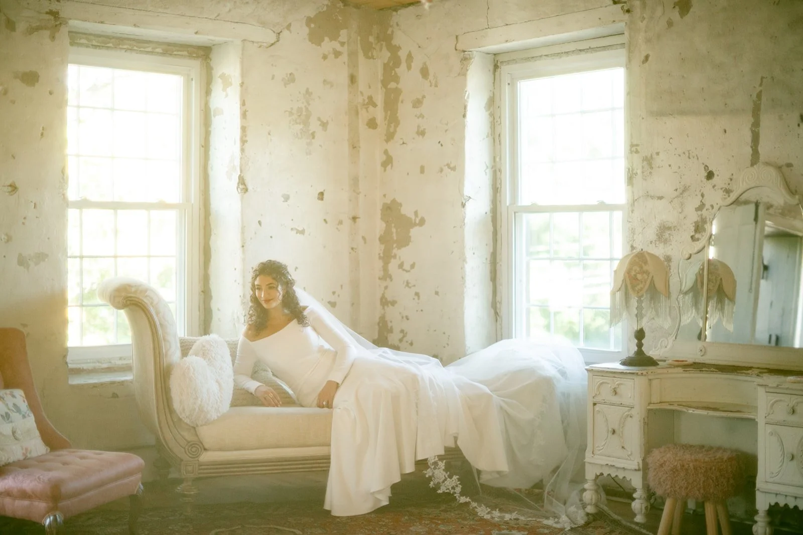 A woman in a white dress reclines on a chaise lounge in a rustic, sunlit room with peeling walls and large windows.