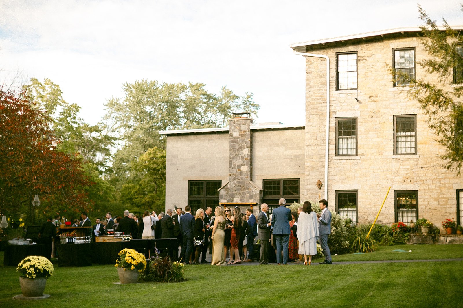 A group of people gathers outside a stone building on a lawn, with tables and potted plants nearby, during a daytime event.
