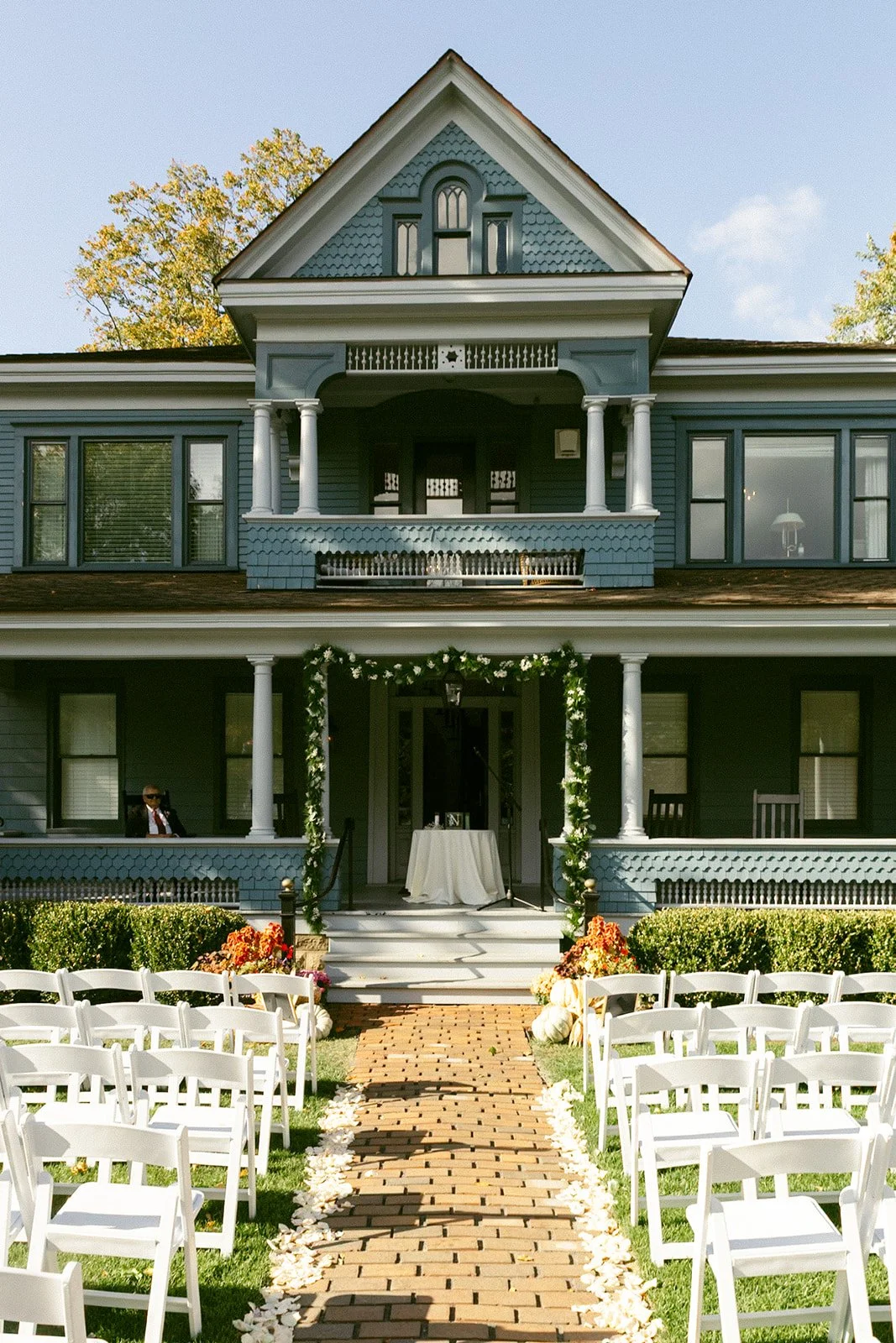 Rows of white chairs face a brick aisle leading to a decorated porch of a large blue house, set up for an outdoor wedding ceremony on a sunny day.