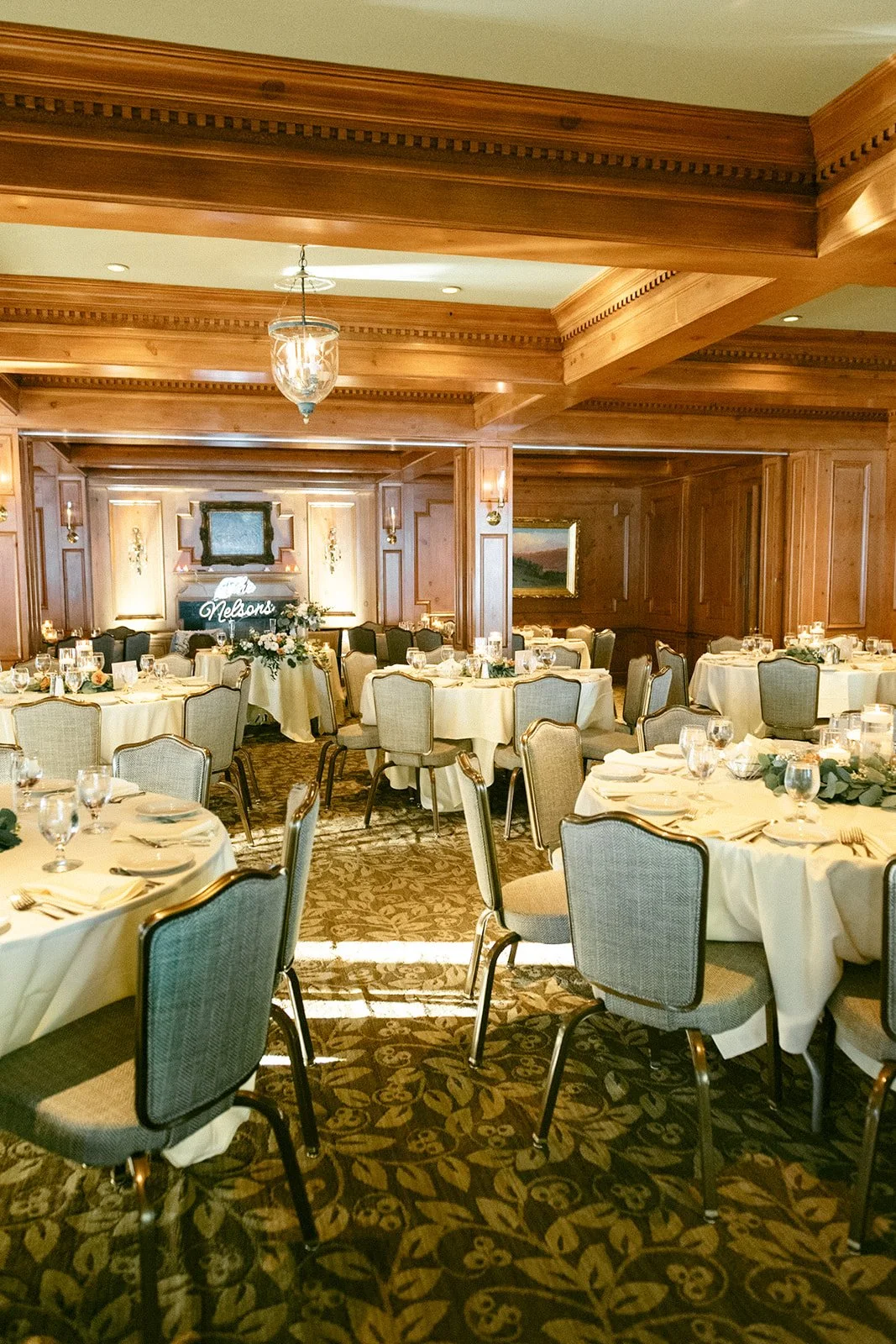 A long dining table set with white linens, glassware, plates, candles, and floral centerpieces in a sunlit room with large windows.