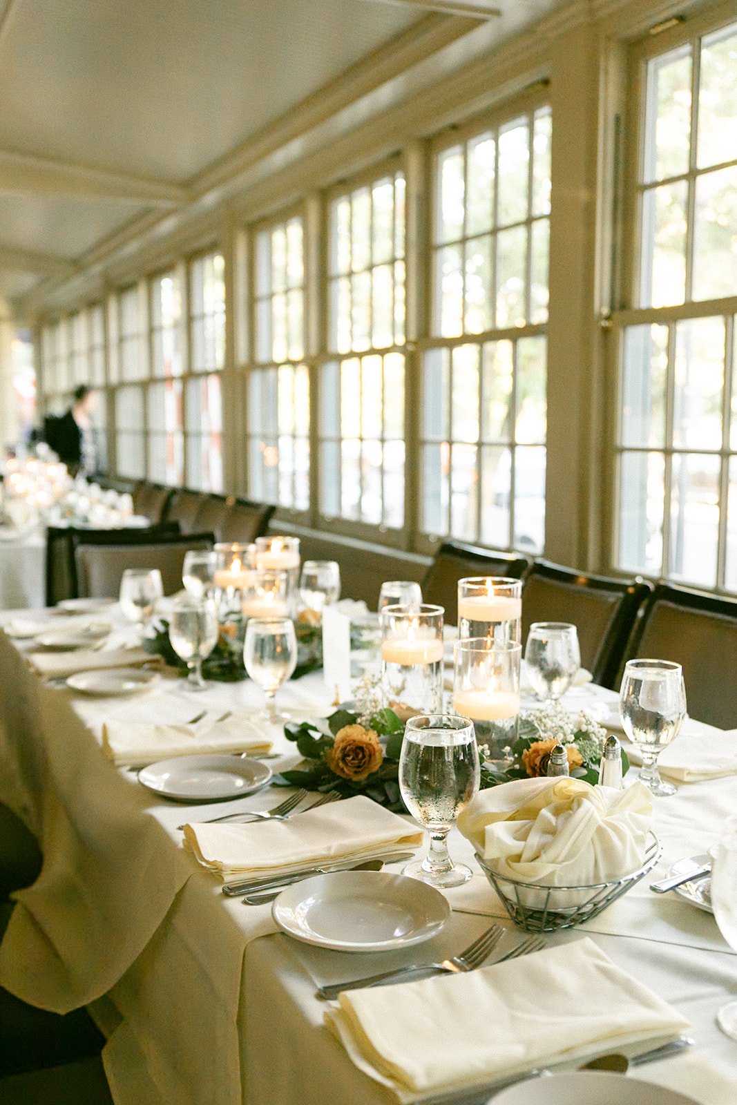 A long dining table set with white linens, glassware, plates, candles, and floral centerpieces in a sunlit room with large windows.
