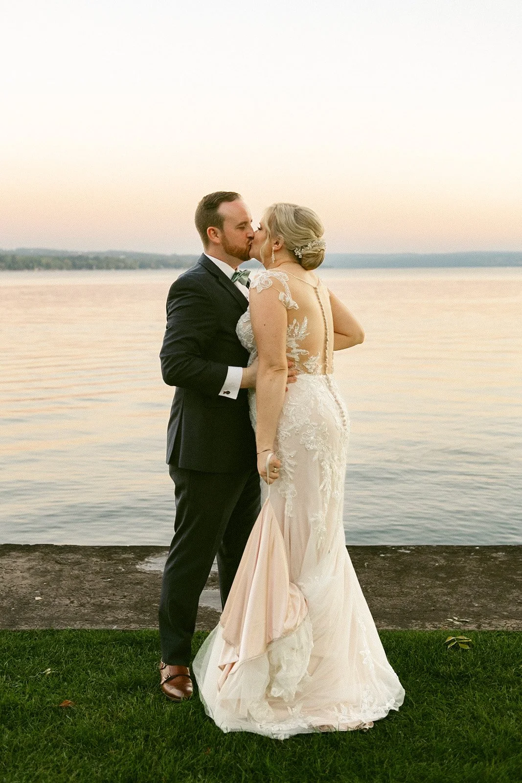 A bride and groom stand facing each other on a grassy edge by a calm lake at sunset, with houses and trees visible in the distance