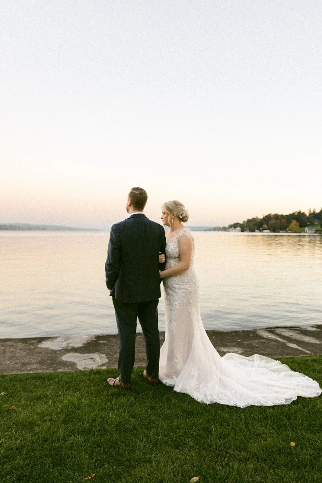 A bride and groom stand facing each other on a grassy edge by a calm lake at sunset, with houses and trees visible in the distance.