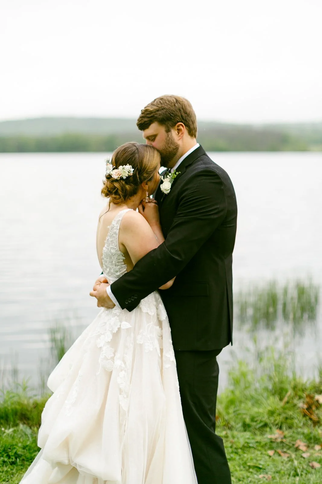 Bride and groom embrace by a lakeside, with the bride in a white gown and the groom in a black suit, standing on grass near the water.