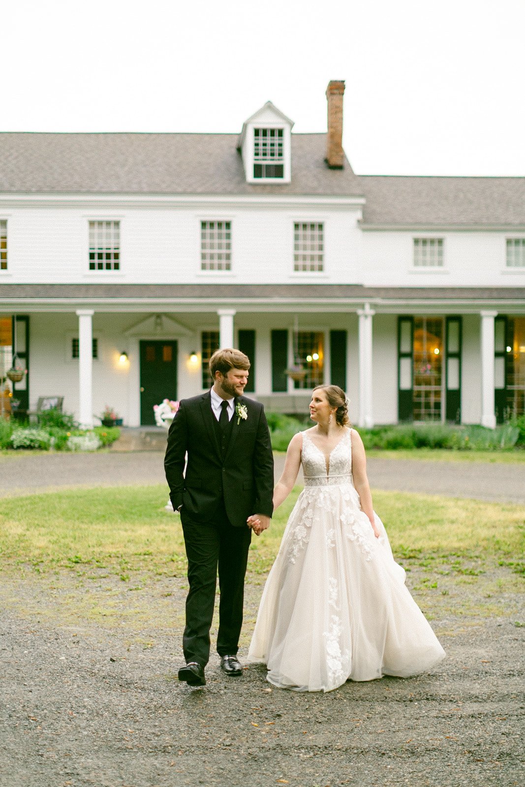 A couple dressed in wedding attire embraces in front of a large, white, two-story house with a porch and several windows.