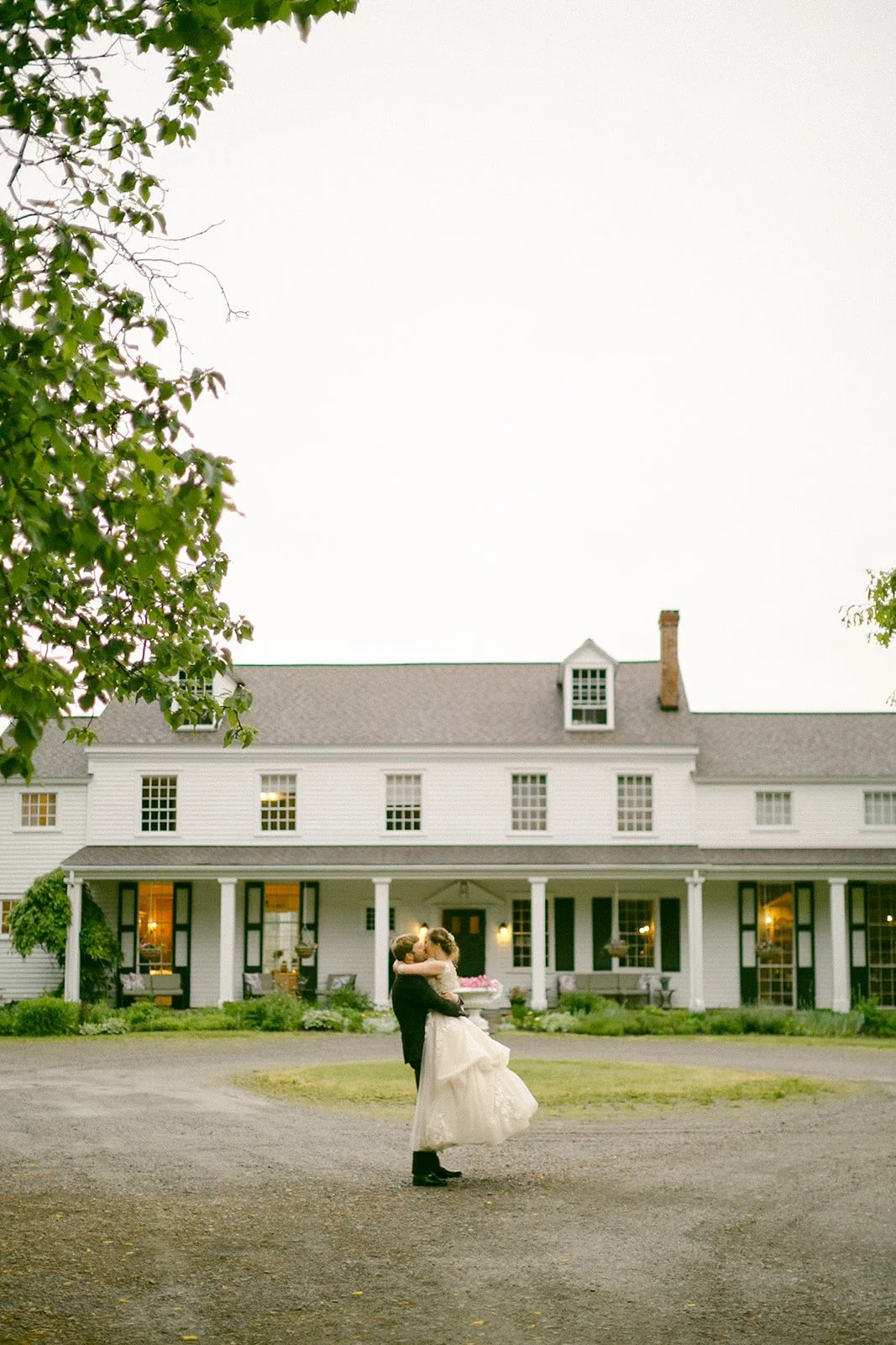 A couple dressed in wedding attire embraces in front of a large, white, two-story house with a porch and several windows.