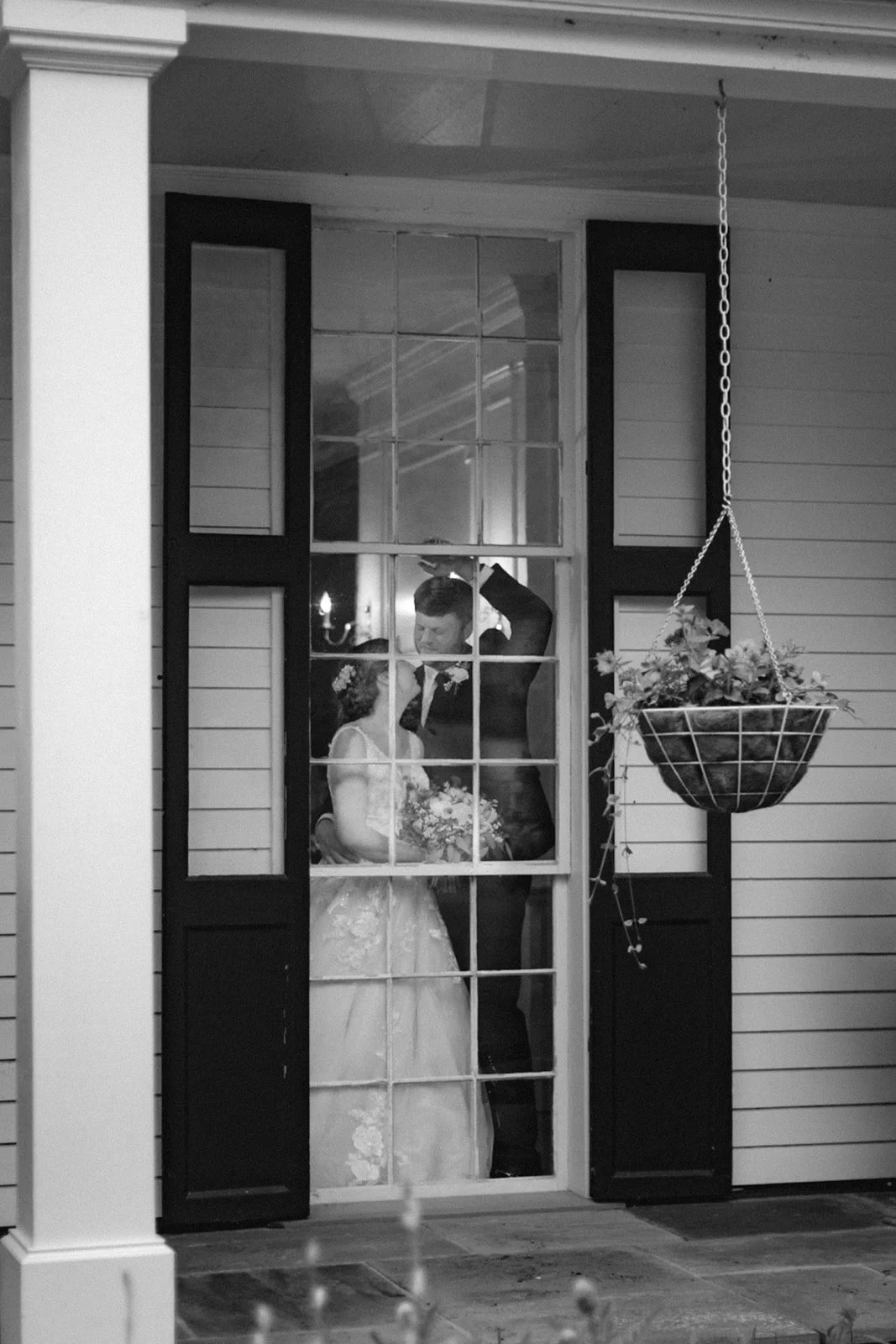 A bride and groom stand together inside a house, visible through glass-paneled doors, with a hanging flower basket in the foreground.