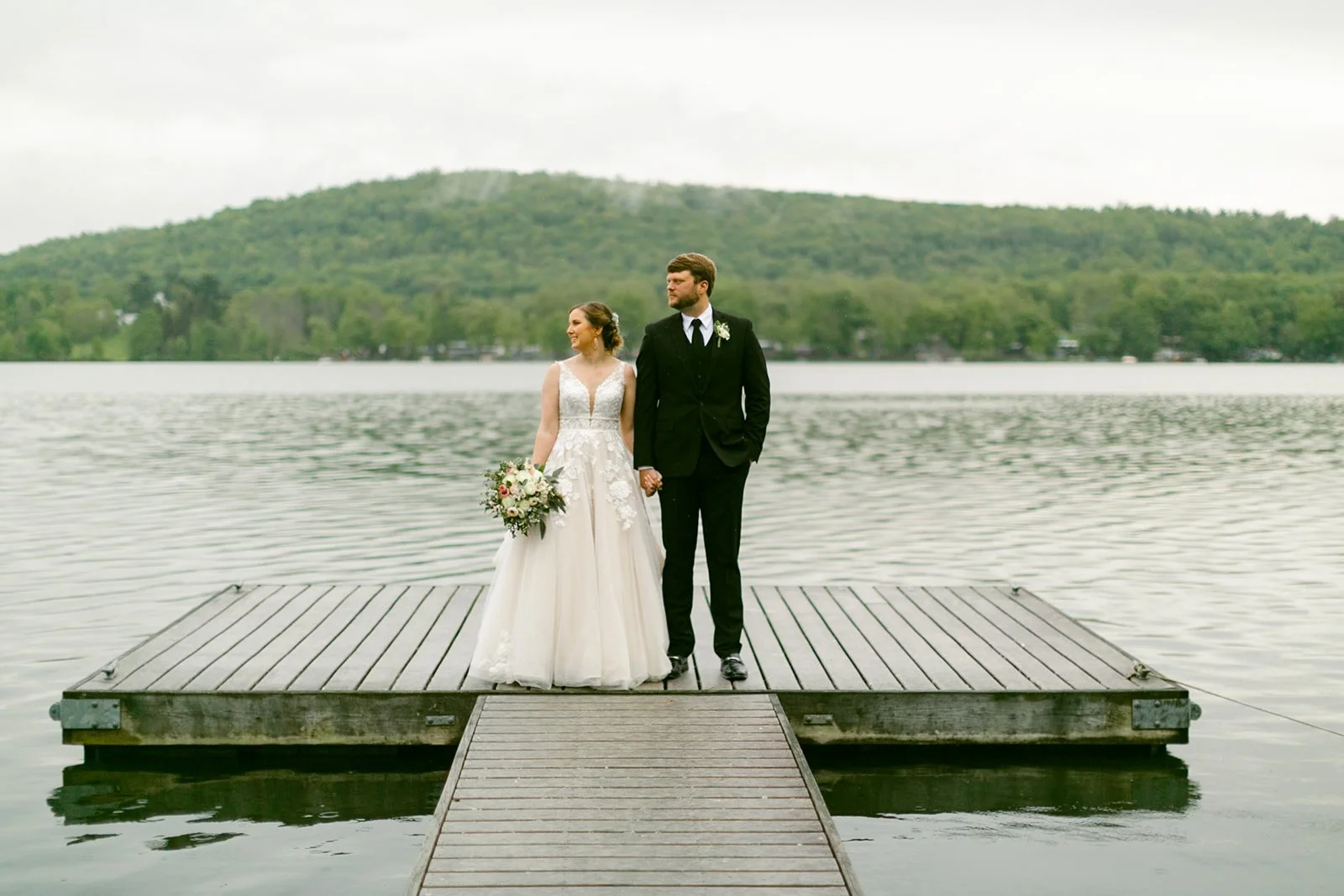 A bride and groom stand on a wooden dock over a lake, holding hands, with a forested hill in the background.