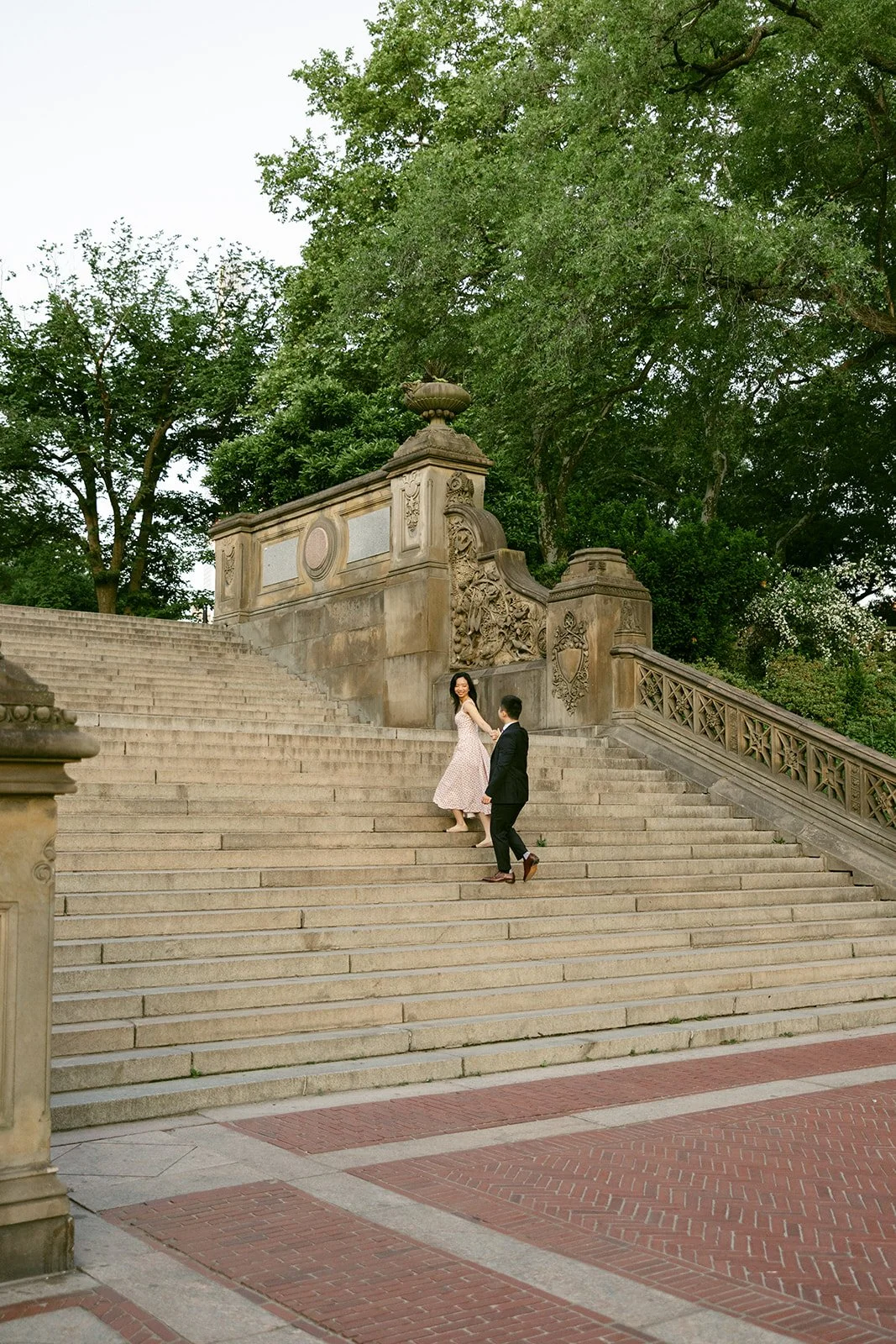 A woman in a light dress and a man in a dark suit walk up wide stone steps outdoors, surrounded by trees and ornate stone railings for central park engagement photos