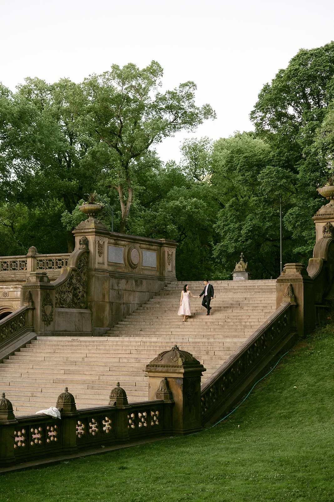 A woman in a light dress and a man in a dark suit walk up wide stone steps outdoors, surrounded by trees and ornate stone railings for central park engagement photos