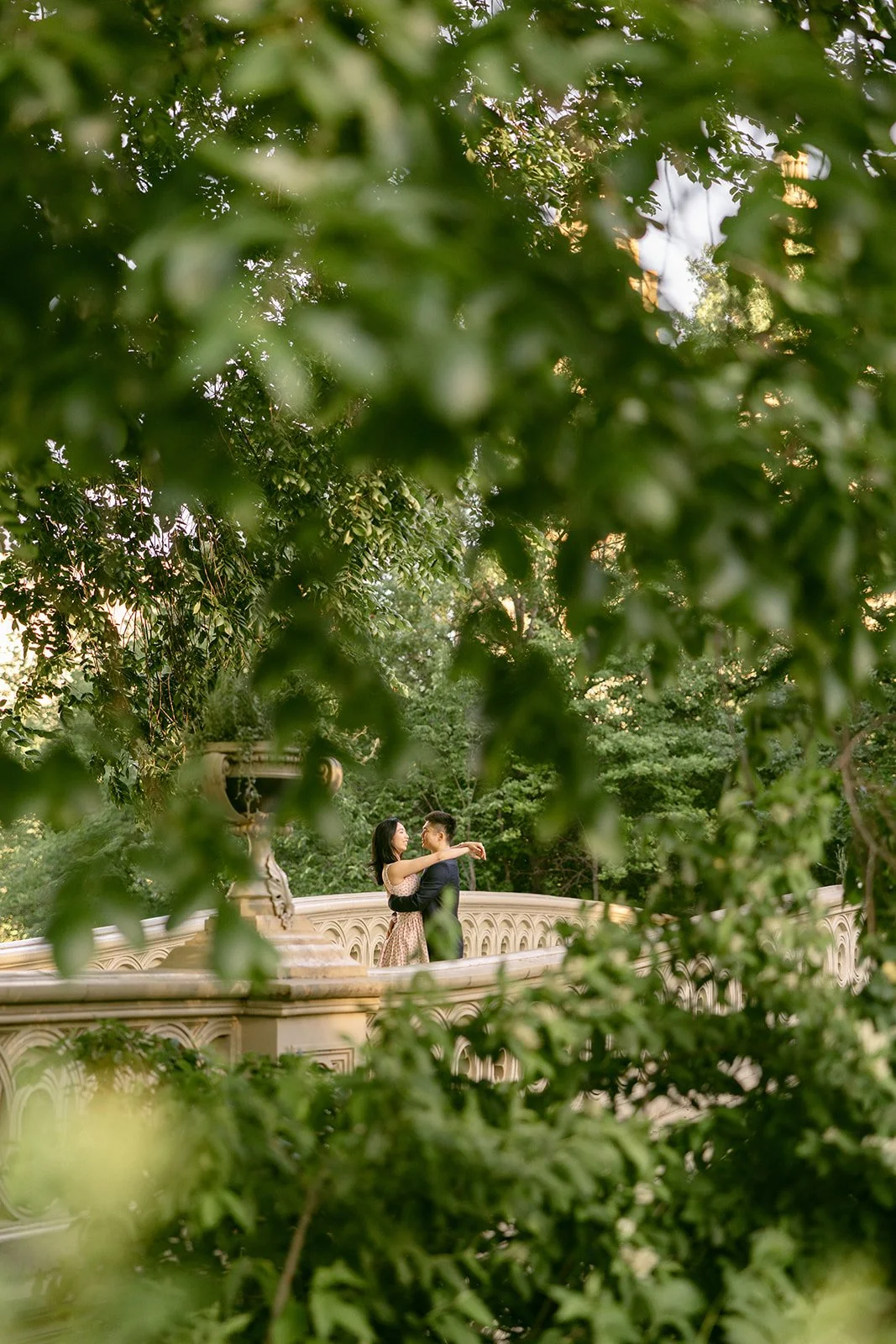 A couple stands close together on a stone bridge, partially obscured by green leafy trees and branches for central park engagement photos