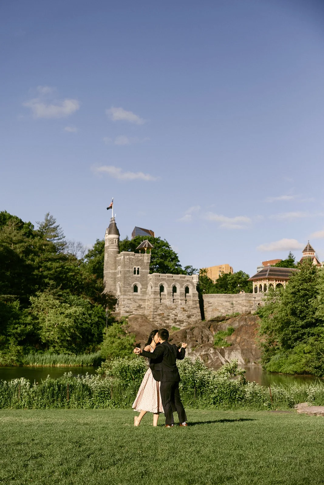 A couple dances on a grassy field in front of a stone castle-like building surrounded by trees under a clear blue sky for central park engagement photos