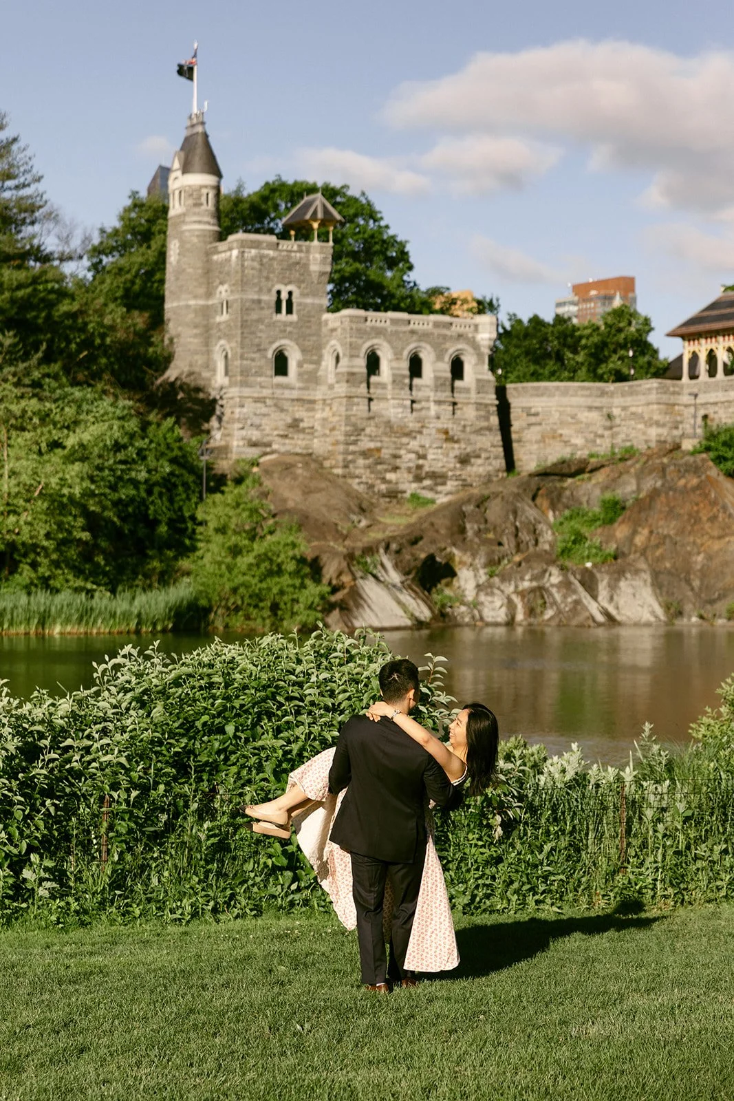 A couple dances on a grassy field in front of a stone castle-like building surrounded by trees under a clear blue sky for central park engagement photos