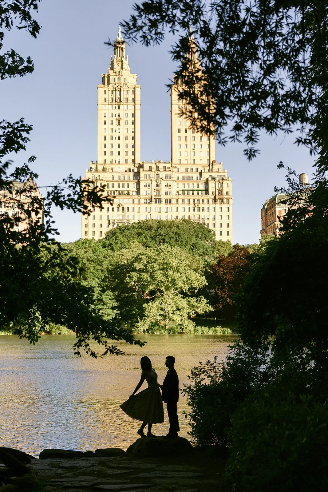 A silhouetted couple stands by a lake in Central Park, with tall buildings and greenery visible in the background on a sunny day.