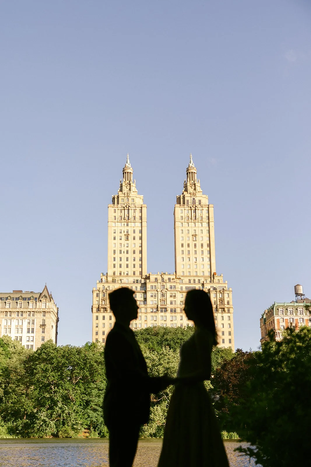 Two people sit by a lake in Central Park, New York City, framed by trees, with the twin towers of the San Remo building in the background for central park engagement photos 
