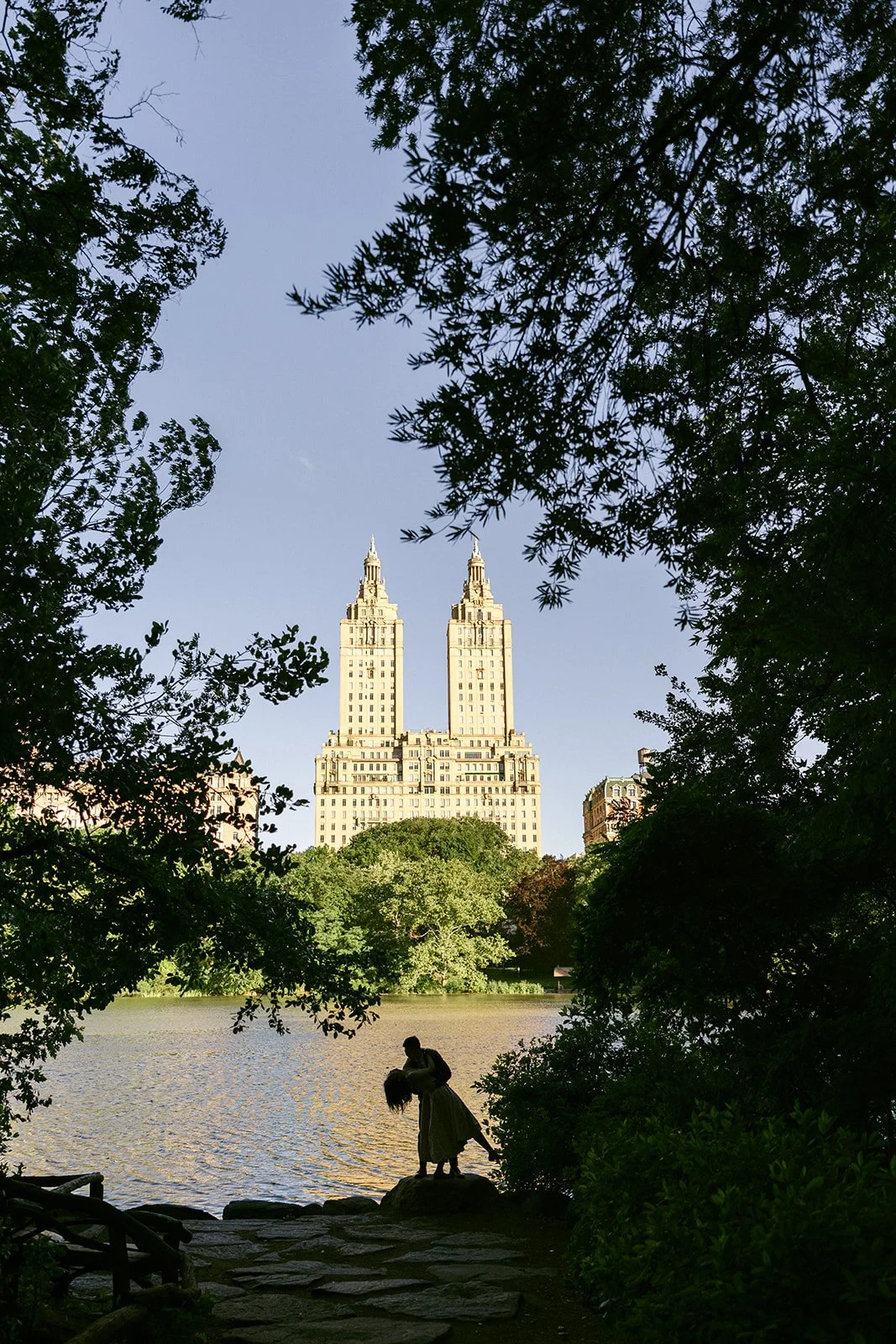 Two people sit by a lake in Central Park, New York City, framed by trees, with the twin towers of the San Remo building in the background for central park engagement photos 