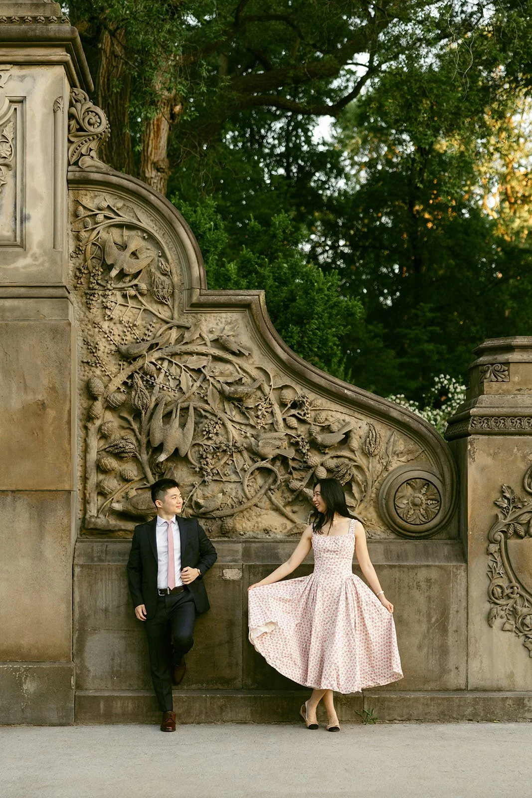 A man in a suit and a woman in a pink dress stand near an ornate stone wall with intricate carvings in an outdoor park setting.