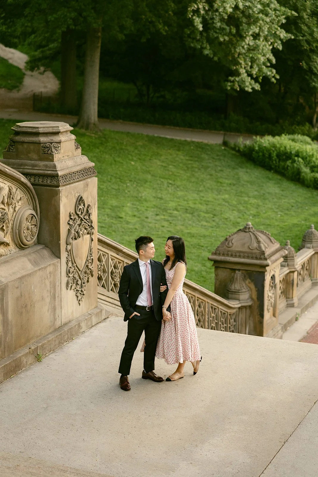 A woman in a light pink dress and a man in a dark suit walk up an ornate stone staircase outdoors, surrounded by trees and detailed carvings for central park engagement photos