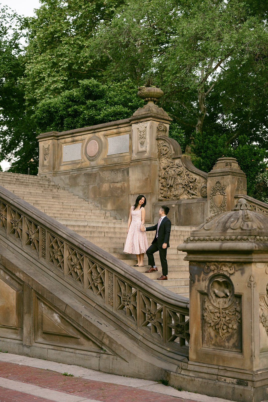 A woman in a light pink dress and a man in a dark suit walk up an ornate stone staircase outdoors, surrounded by trees and detailed carvings for central park engagement photos