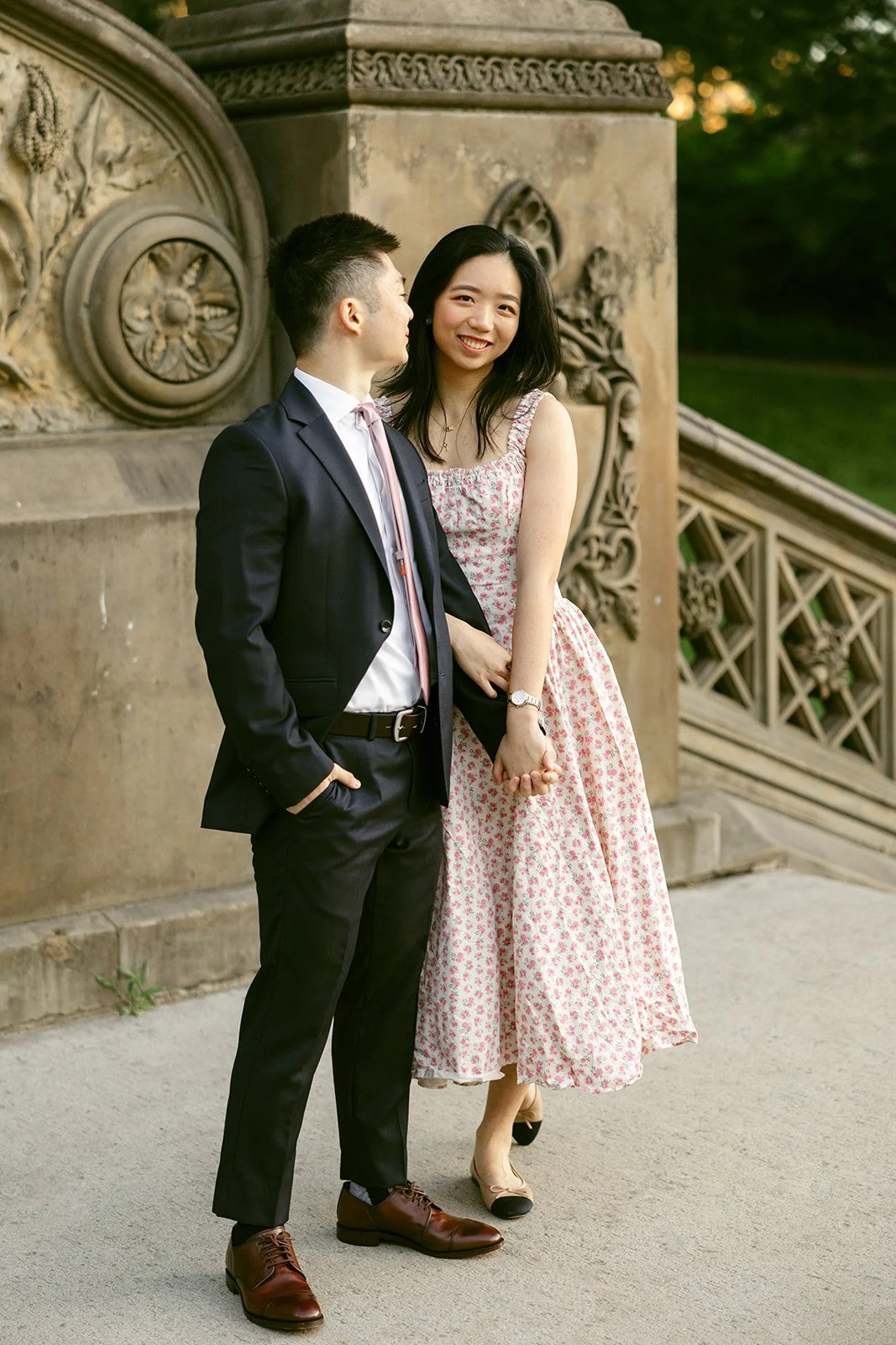 A woman in a light pink dress and a man in a dark suit walk up an ornate stone staircase outdoors, surrounded by trees and detailed carvings for central park engagement photos