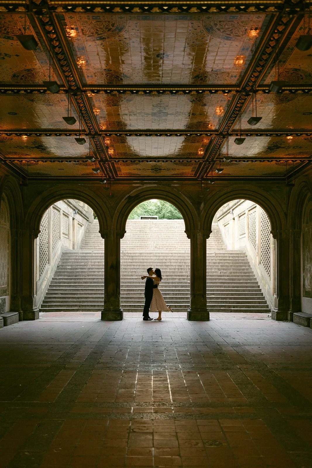 A couple stands embracing beneath an ornate, arched ceiling in a covered walkway, with steps and greenery visible in the background.