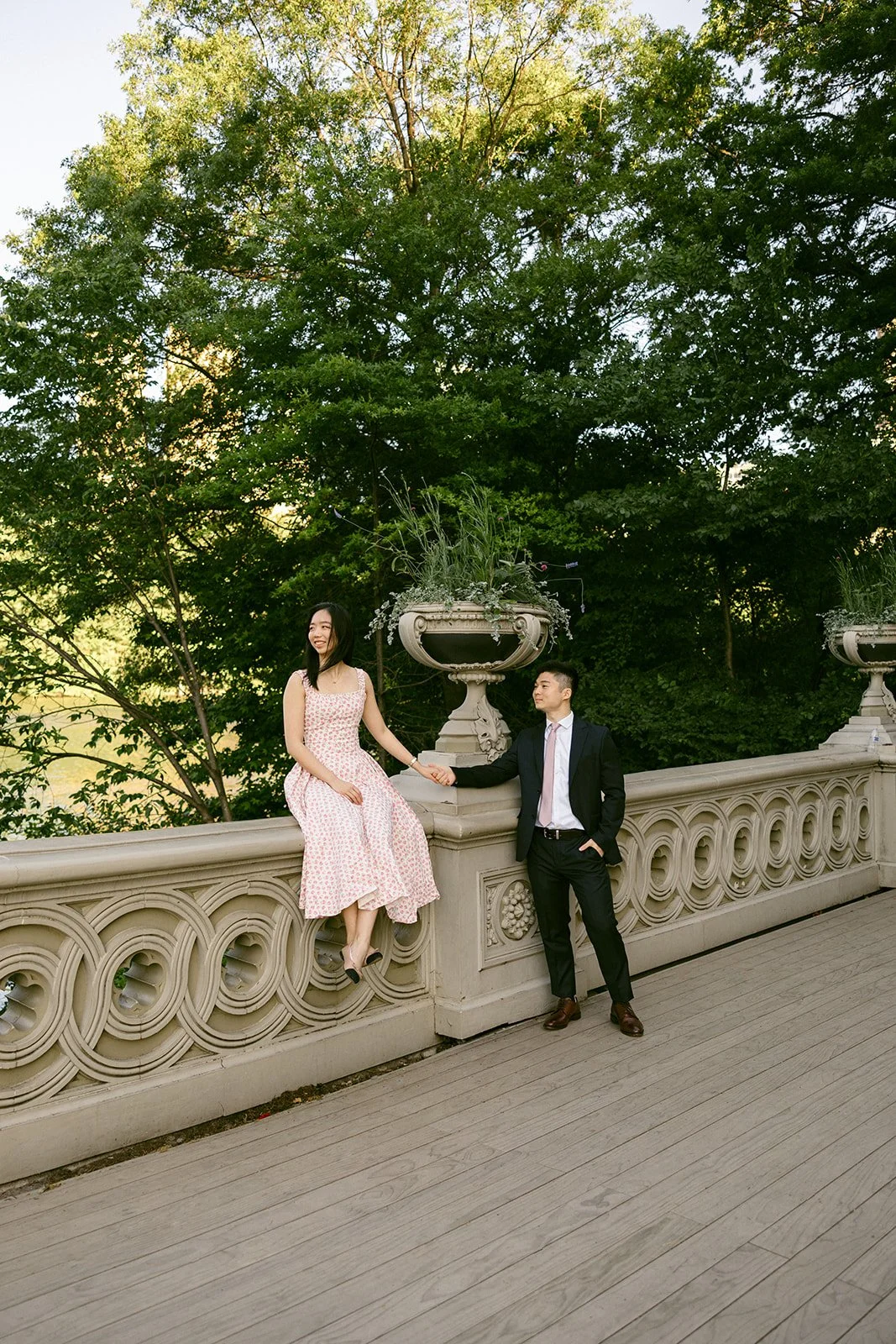 A woman in a light dress sits on a decorative stone railing while a man in a suit stands nearby; both are outdoors surrounded by trees and greenery.