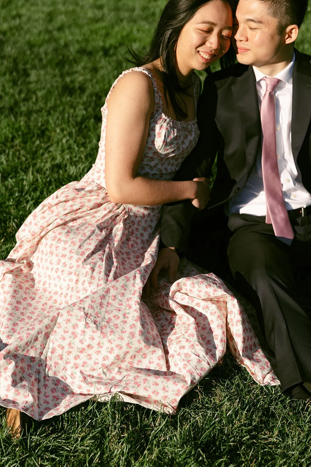 A woman in a floral dress and a man in a suit sit closely together on grass, both smiling and leaning towards each other.