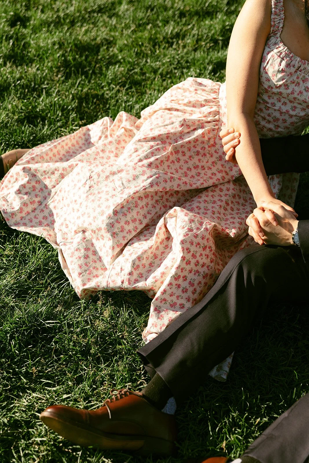 A woman in a floral dress and a man in a suit sit closely together on grass, both smiling and leaning towards each other.