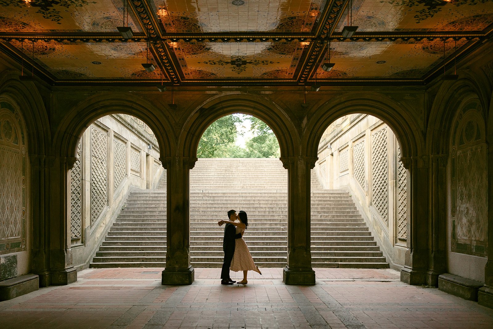  A couple stands under an ornate archway, embracing, with wide stone steps and greenery visible in the background.